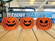 Three orange powder-coated steel jack-o'-lanterns with unique carved faces standing in a row on a wooden surface, showing a dual-layer 3D design.