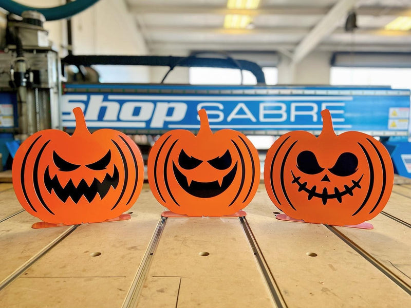 Three orange powder-coated steel jack-o'-lanterns with unique carved faces standing in a row on a wooden surface, showing a dual-layer 3D design.