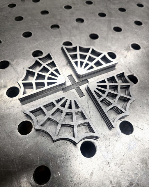 Two 5-inch spider web decorative steel gussets placed on a metal fabrication welding table with circular mounting holes.